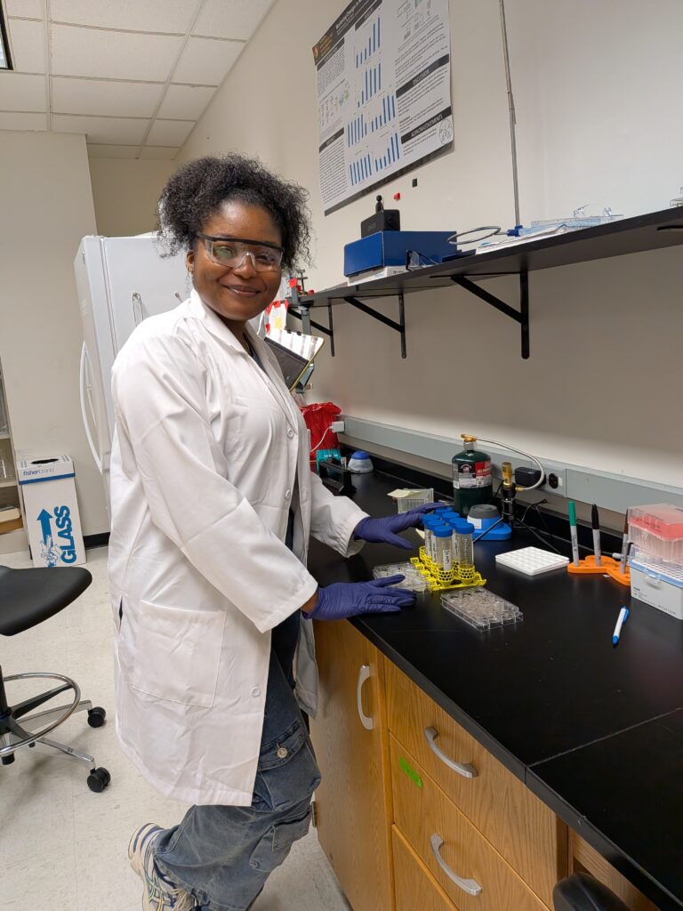 A student researcher in a lab coat, safety goggles, and gloves works at a lab bench with test tubes and scientific equipment.