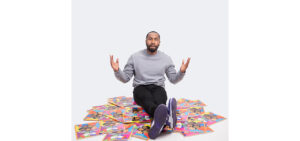 A person sits on the floor with hands raised, surrounded by numerous copies of a colorful, illustrated book cover.