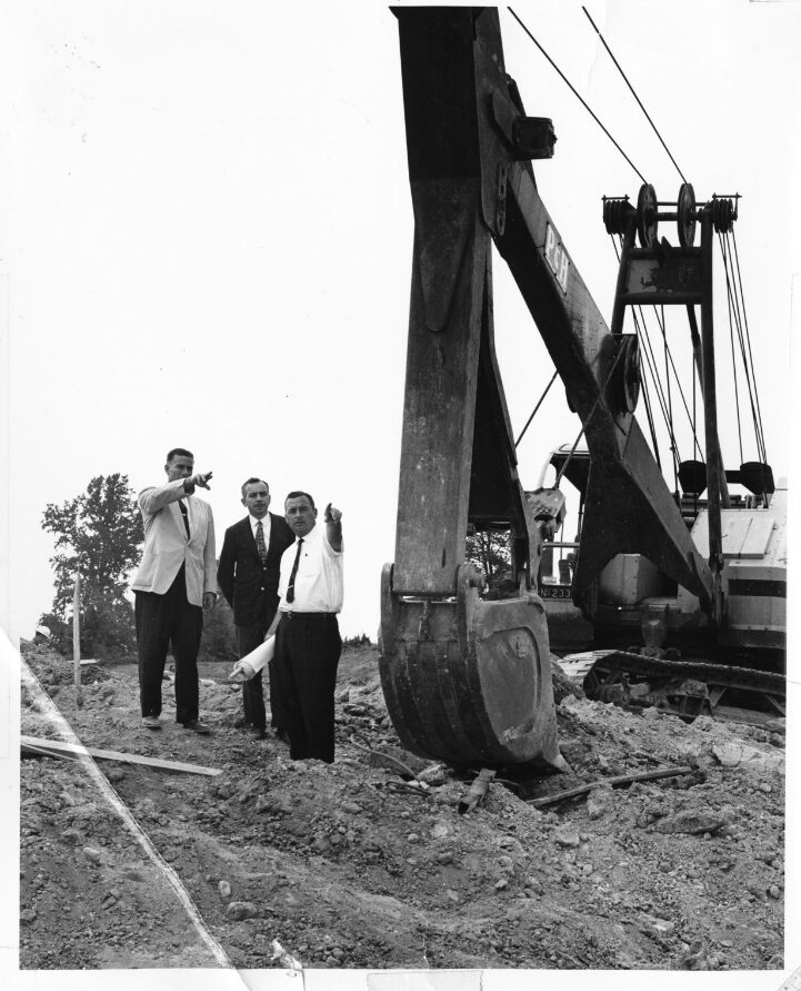 Black-and-white photo of three men in suits pointing across an excavated construction site beside a large power shovel