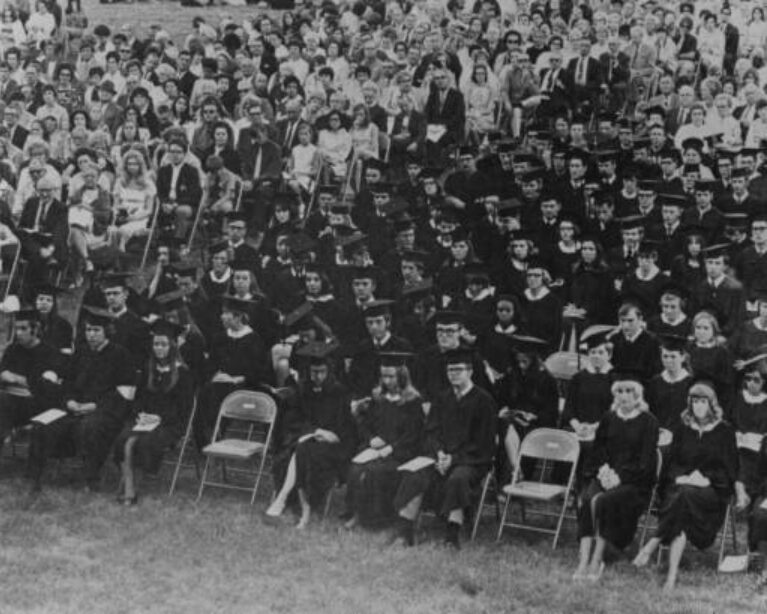 Black-and-white photo of graduates in caps and gowns seated on a lawn in front of a large crowd of family and guests