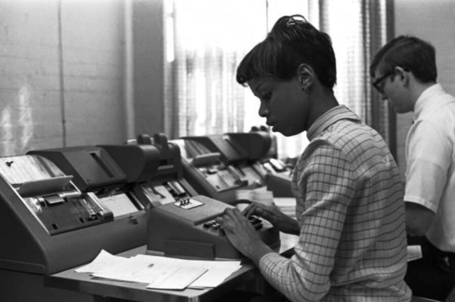 Black-and-white photo of two students working at rows of early data-processing machines