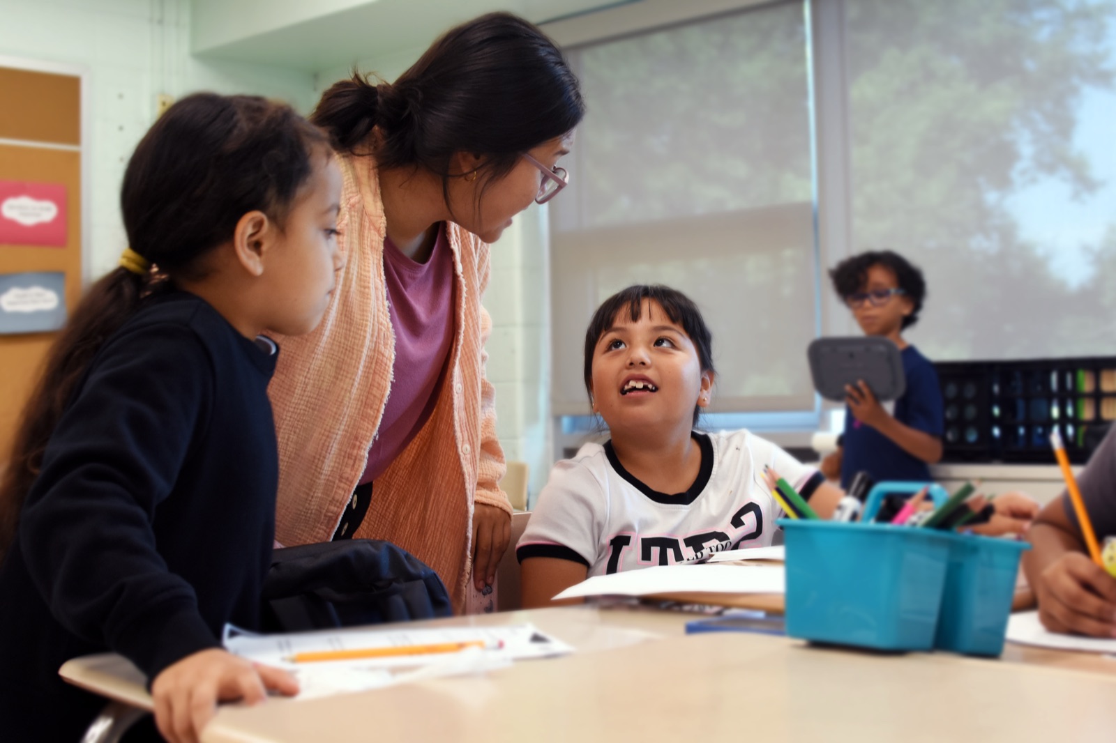 An instructor leans over to talk with two students at a desk, while another student stands in the background holding a tablet.