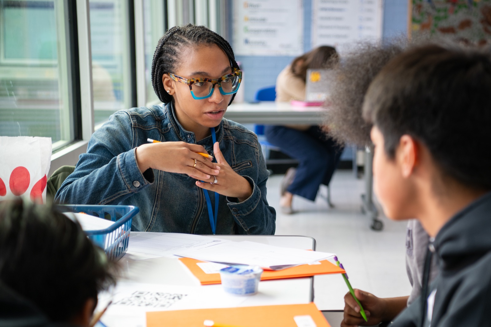 A person talks to students at a table while gesturing with a pencil. Documents and an orange folder are on the table.