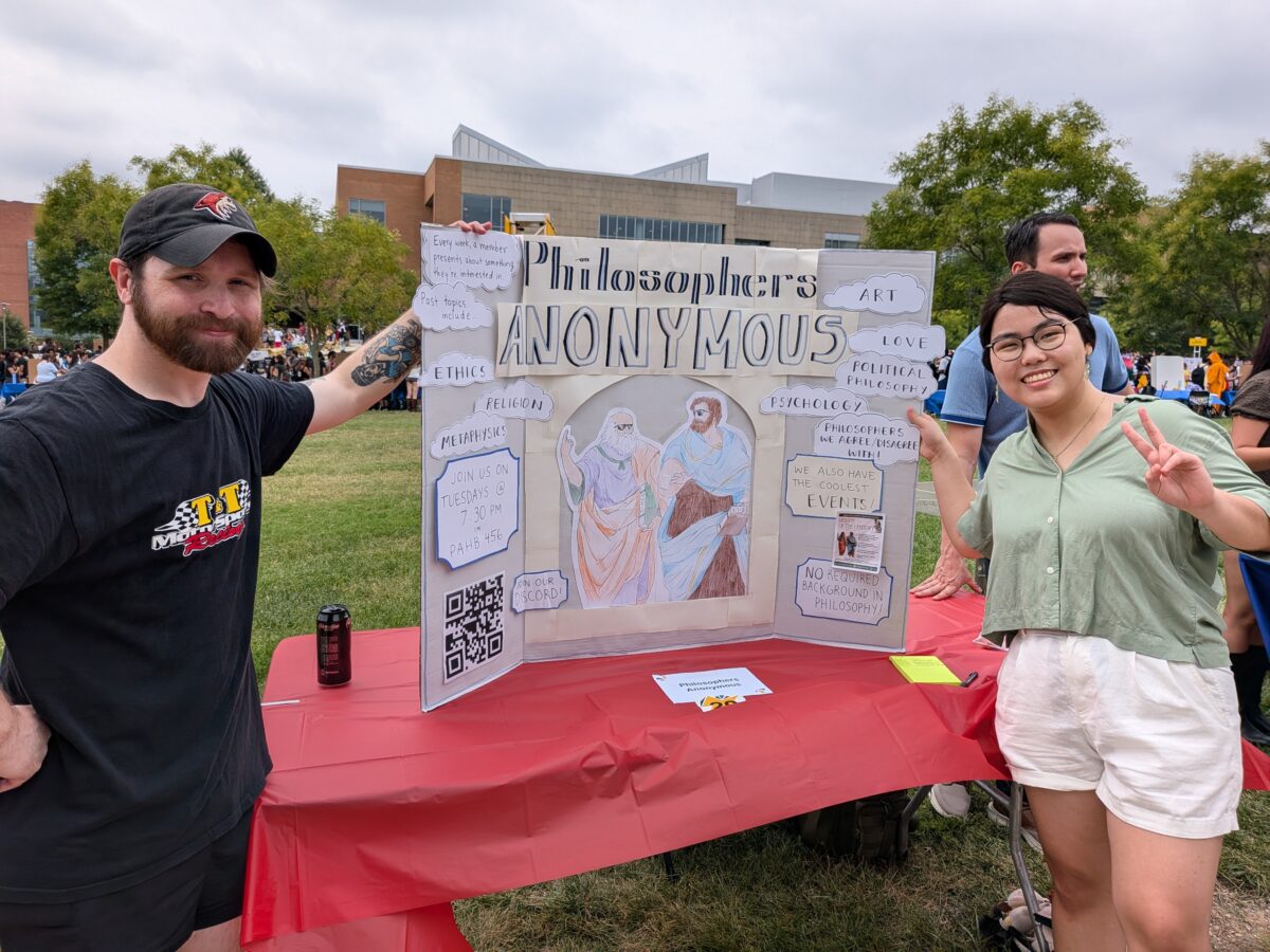 Two college students stand on both sides of a presentation board that shows information about a philosophy club