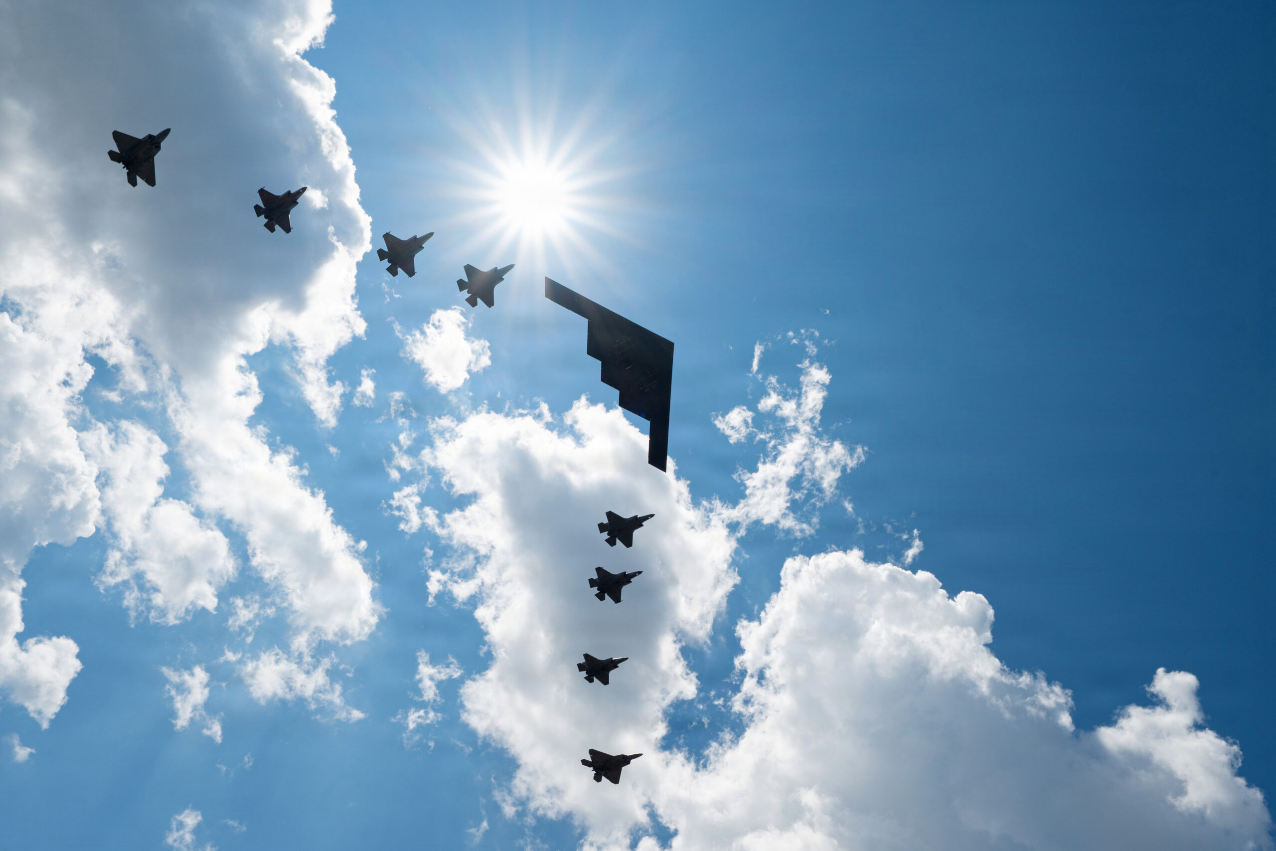 one large air force aircraft in formation with seven smaller aircraft, viewed from below, with bright blue sky and puffy white clouds in the background
