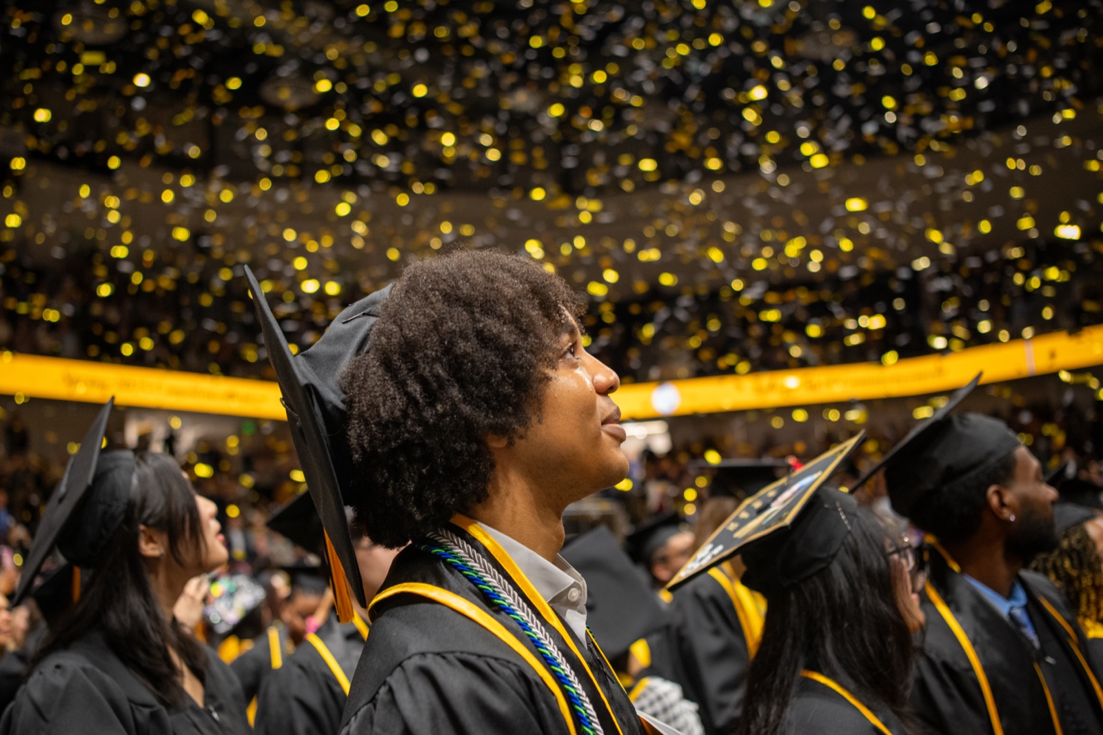 A UMBC graduate in cap, gown, and honor cords looks up during commencement as gold confetti falls