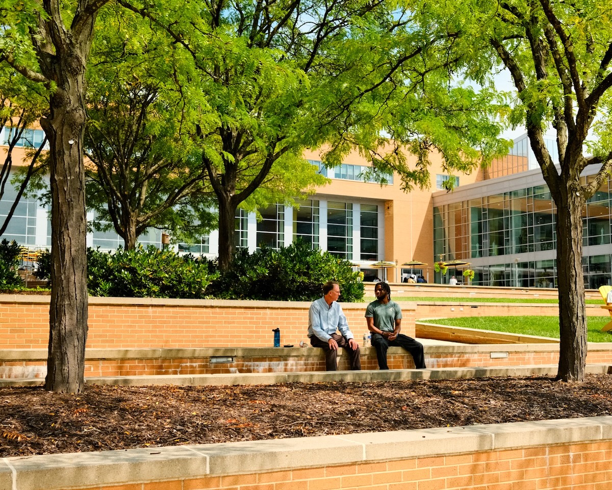 Two people sit on a low brick wall outside on campus, talking to each other.