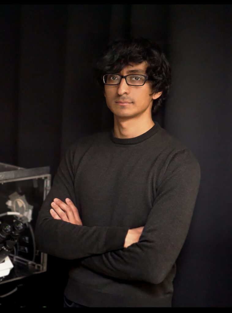 Headshot of a person with dark, wavy hair and glasses, wearing a dark sweater with arms crossed.