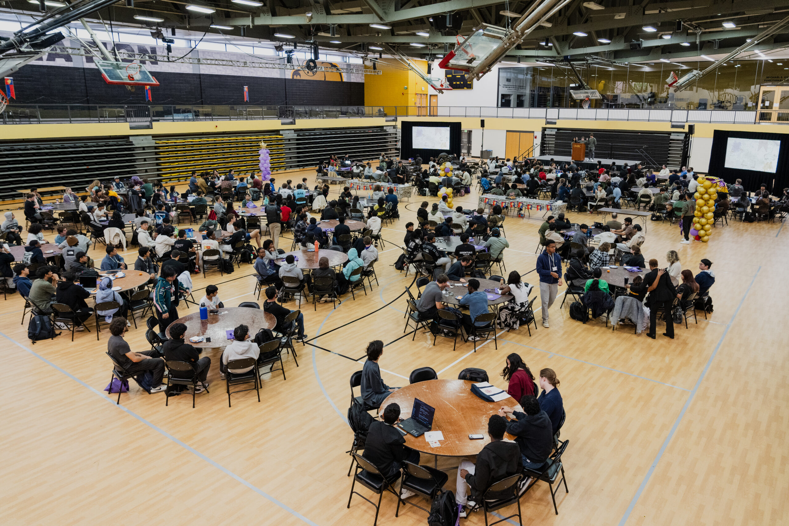 Students gather at round tables to start hackUMBC, with two projection screens visible at the front of the room.