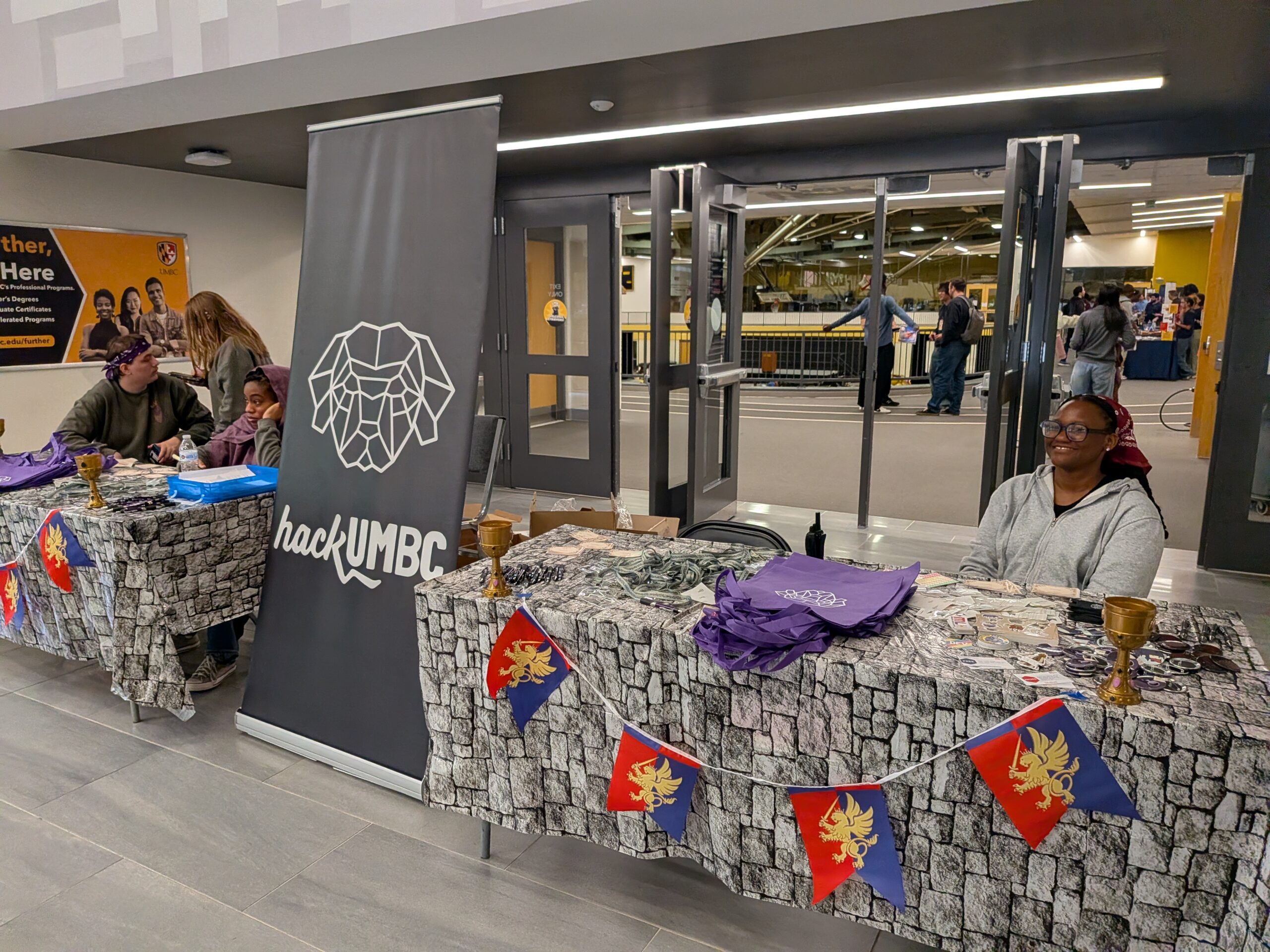 Students sit at tables with stone patterned cloths and banners for hackUMBC. One student sits alone at a table with promotional items.